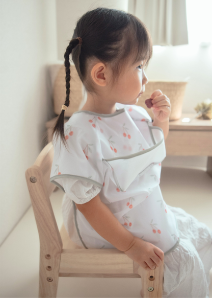 女の子が半袖お食事エプロンを着用して椅子に座っている使用イメージ。身体の前面を覆うデザイン
