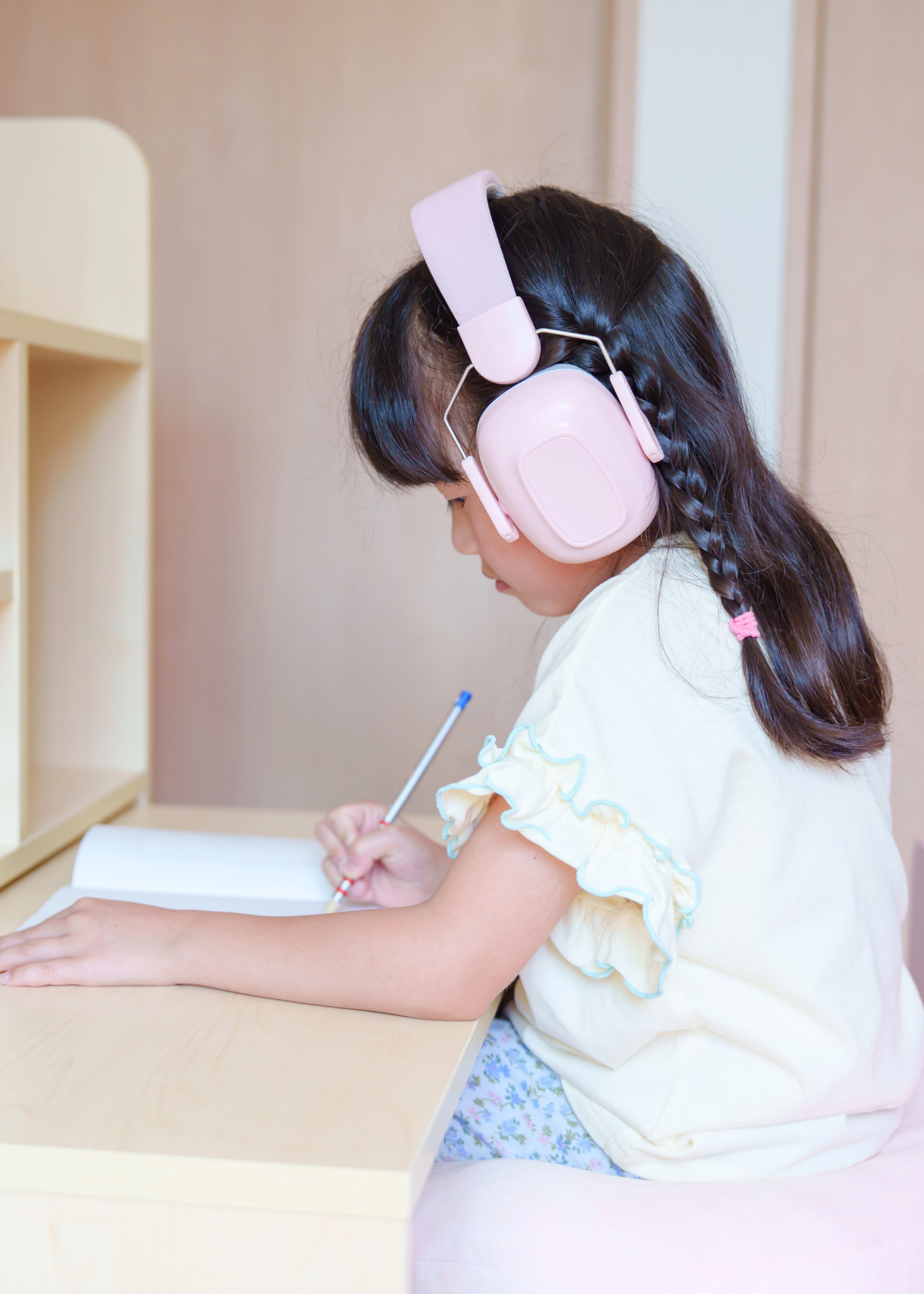 ピンクのキッズ防音イヤーマフを女の子が着用している使用イメージ。机に向かって過ごしている様子