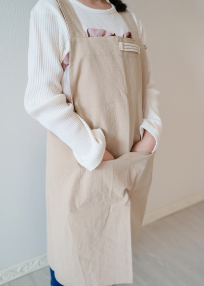 ベージュの親子エプロンを着用した大人の正面イメージ。大きな前ポケットとバッククロス型の肩ひもを確認できるデザイン