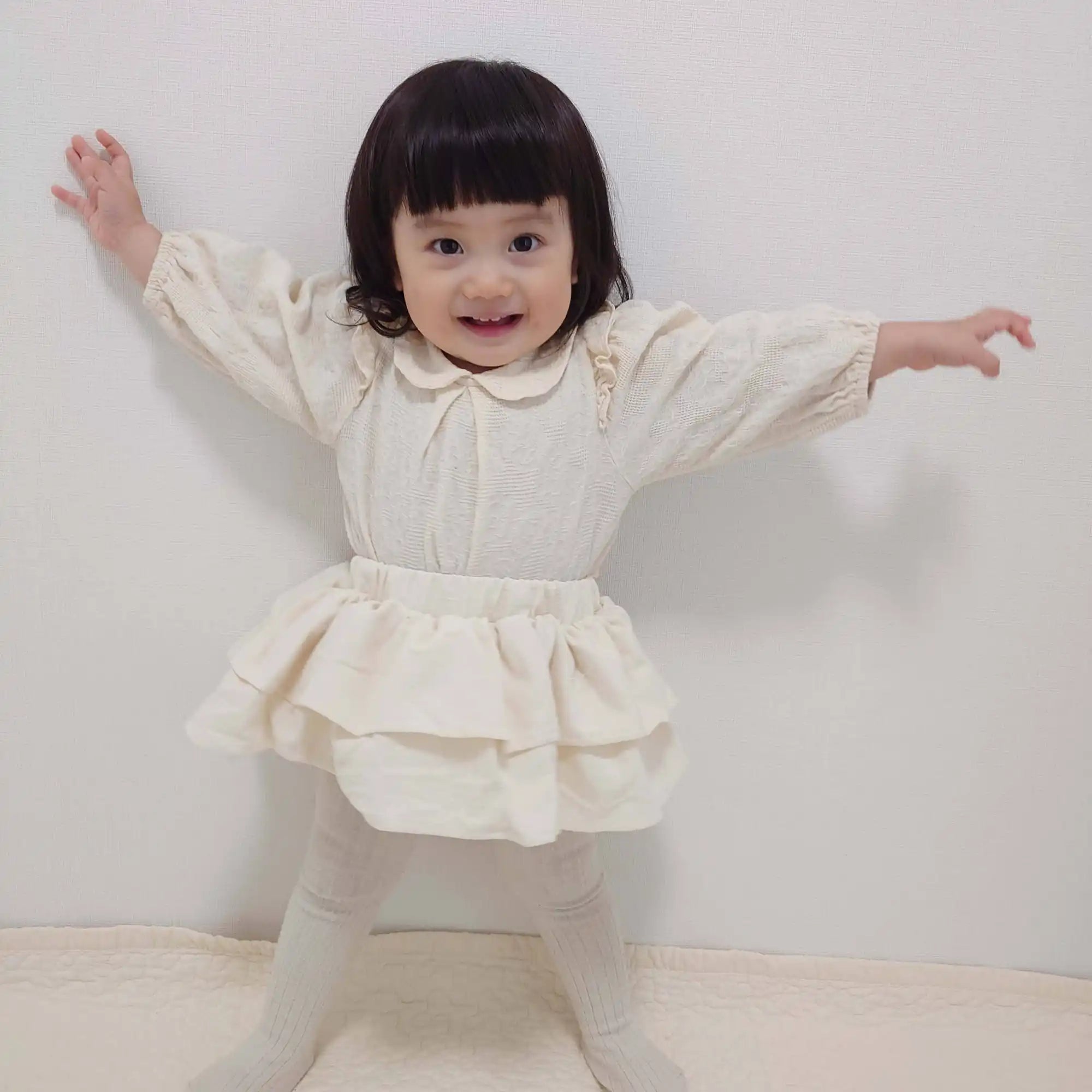レース襟のクリーム色のトップスと、同じ色のティアードスカートとタイツを履いた愛らしい幼児。