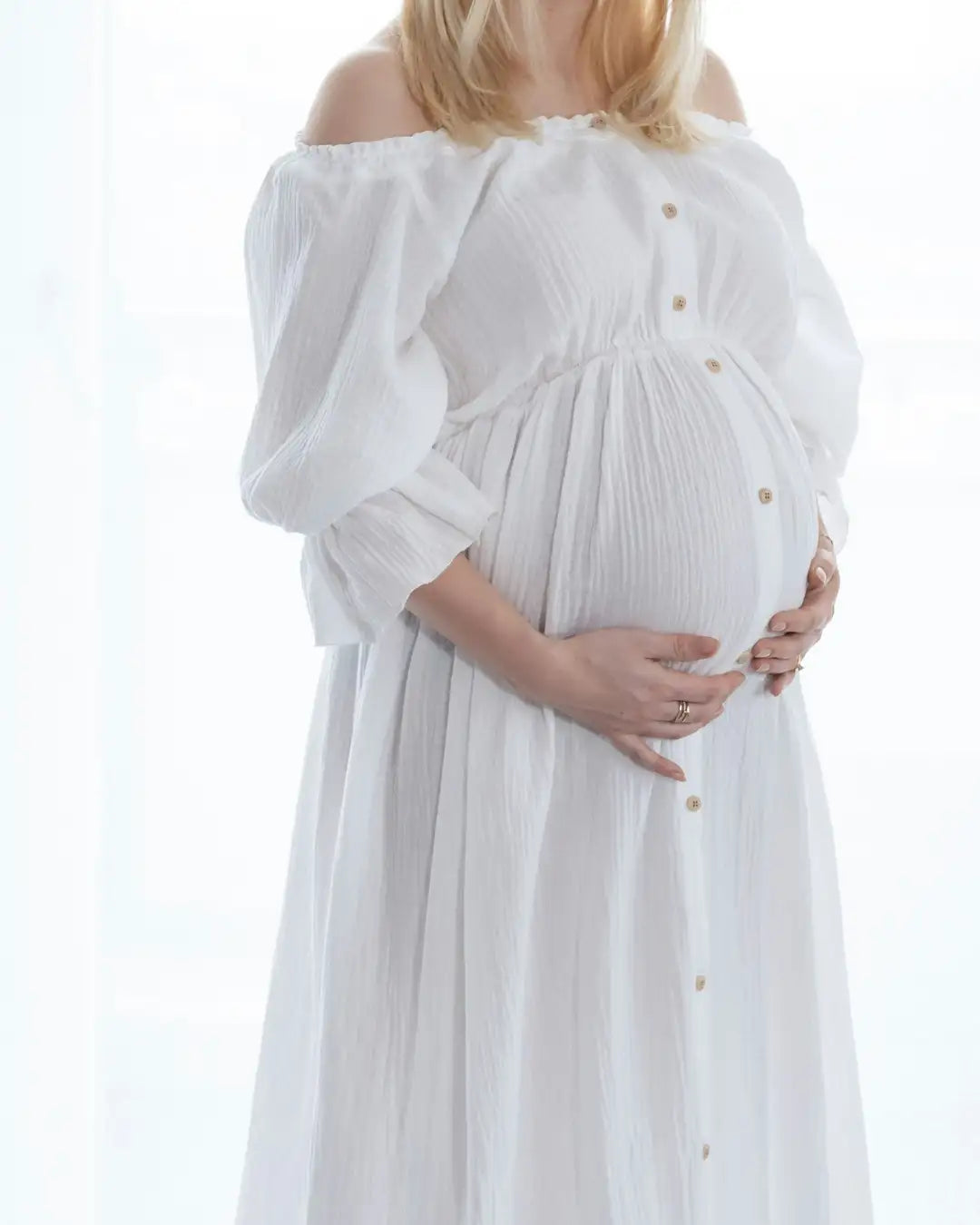 ボタンのディテールと柔らかくギャザーされた生地が特徴の、流れるような白いオフショルダーのマタニティドレス。