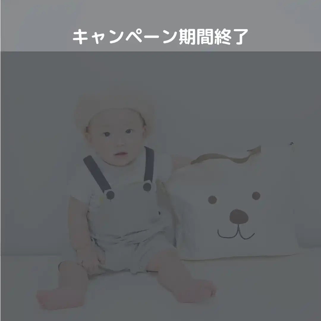 白いオーバーオールと白い帽子をかぶった赤ちゃんが、白いくまの形の玩具箱のそばに座っています。
