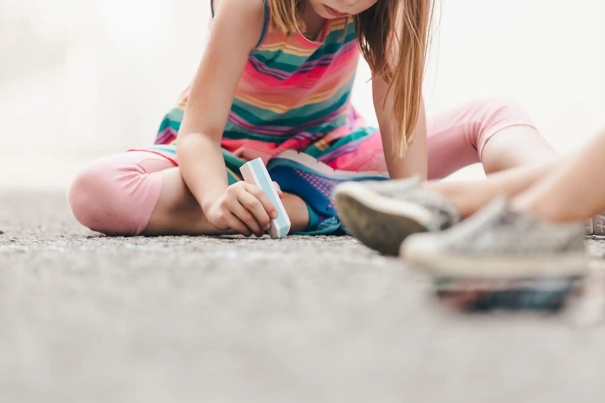 カラフルな縞模様のドレスとピンクのレギンスを着た若い女の子が、地面に淡い青色のチョークで絵を描いています。