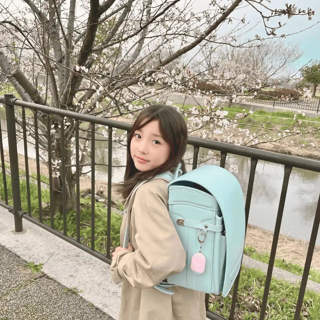 薄い茶色のコートを着て、薄い青色のランドセルを背負った若い女の子。