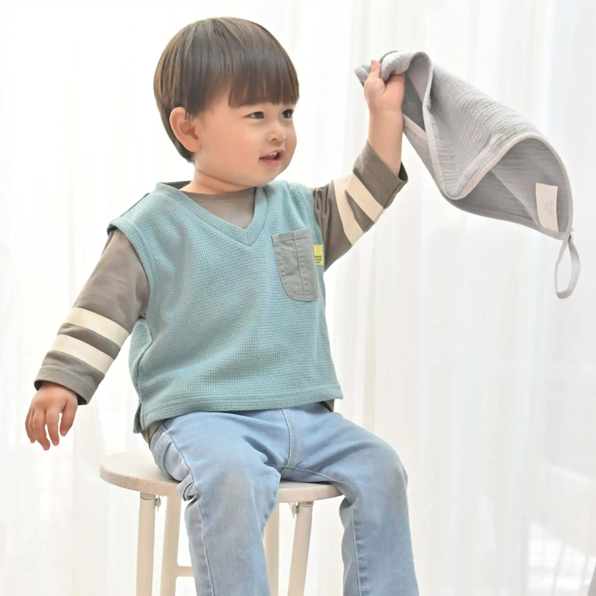 青いベストと薄い青いジーンズを着た幼い子供が、灰色のブランケットを掲げている。