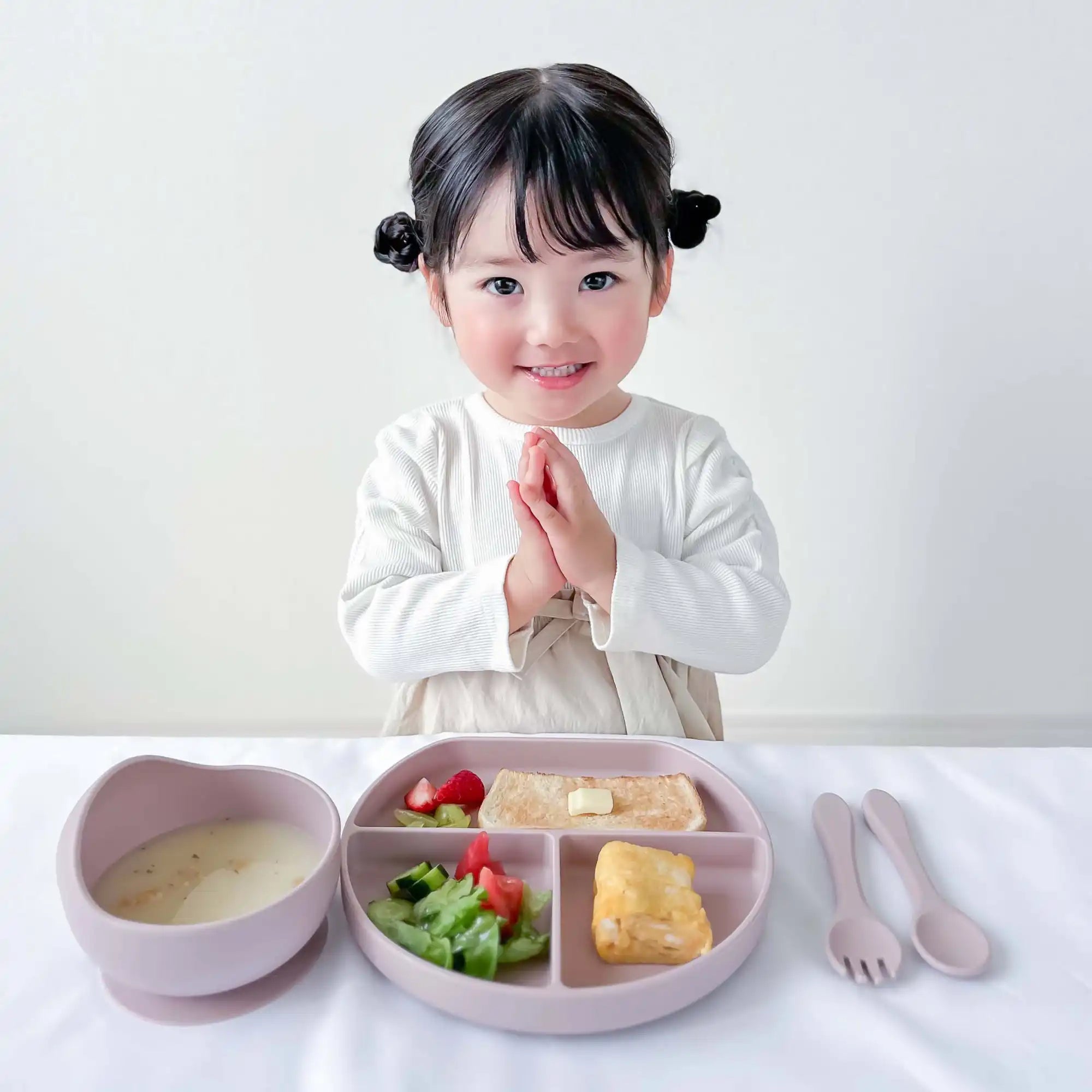 手を合わせて笑っている幼い子供が、仕切られた皿の食べ物とスープの入ったボウルを持ったテーブルに座っている。