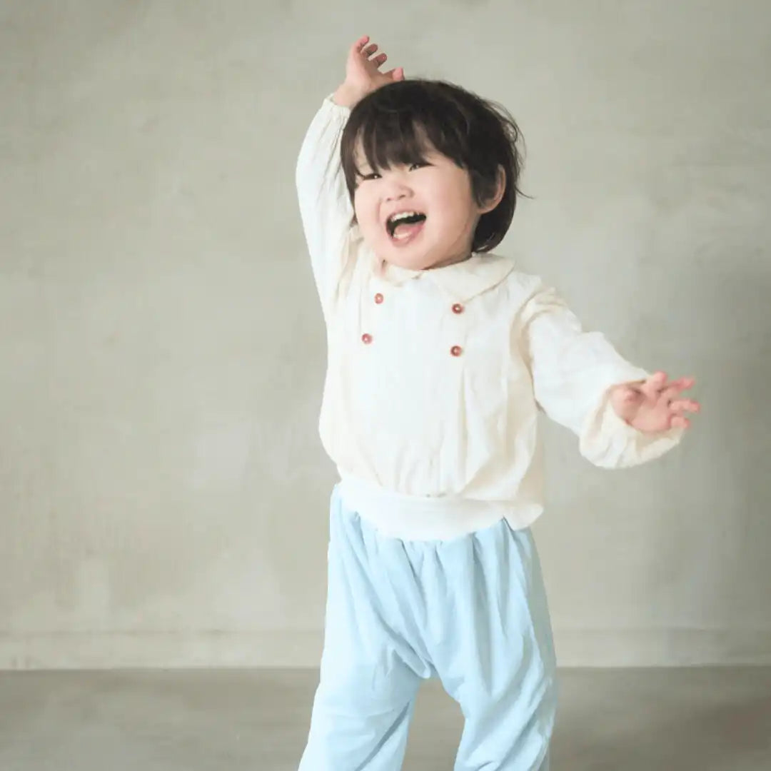 白い長袖シャツに赤いボタンと淡い青色のズボンをはいた子供。